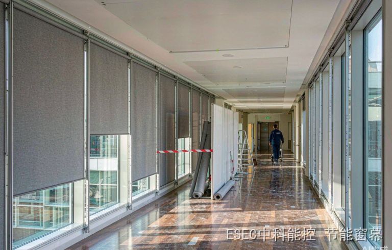 办公楼走廊一侧整排窗已装好灰色遮阳窗幕、部分半降，另一段窗户仍是裸露玻璃，临时隔断与警示带分隔作业区，工人推料车走向电梯