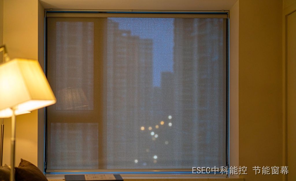 A winter night living room with a light grey translucent screen drawn down in side rails over a floor-to-ceiling window, blurred city lights glowing through the fabric