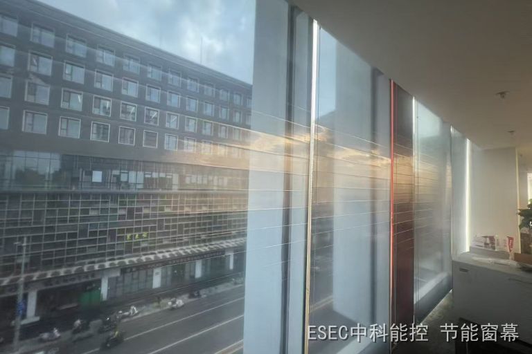 从室内看整排玻璃幕墙，半降的隔热透景窗幕覆盖在玻璃内侧，窗外是待更新的城市街道与沿街建筑，室内工位与绿植保持日常使用状态