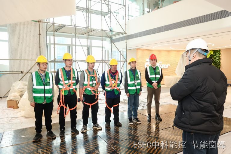 从室内看到的玻璃幕墙更换施工现场，施工班组佩戴安全帽与安全带在脚手架旁集合，背景是大面积玻璃幕墙与室内中庭