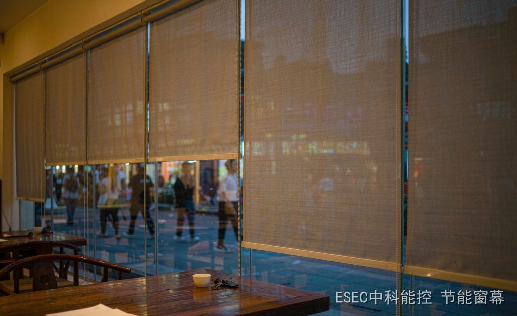 Inside a street-front tea house at dusk, a light grey translucent shading screen is drawn two thirds of the way down in slim side rails on the room side of a glass curtain wall, with a teacup, keys and a folded white notice on the wooden table
